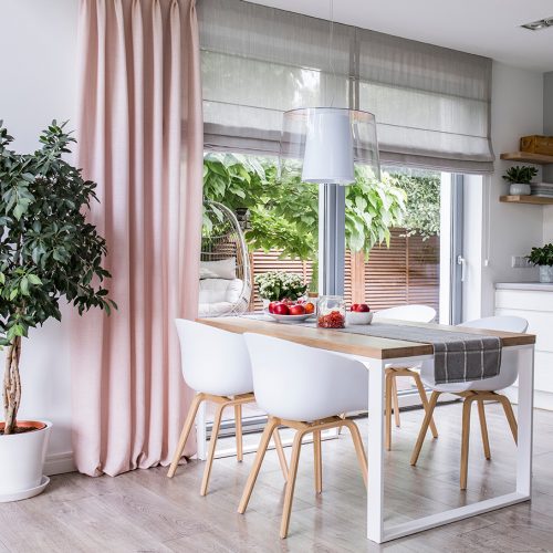 Gray roman shades and a pink curtain on big, glass windows in a modern kitchen and dining room interior with a wooden table and white chairs; Shutterstock ID 1147624868; Purchase Order: -